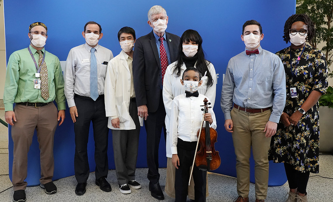 Caesar stands proudly holding violin and bow in front of his NIH physicians and other health staff and the Clinical Center chaplain, with Collins in the middle