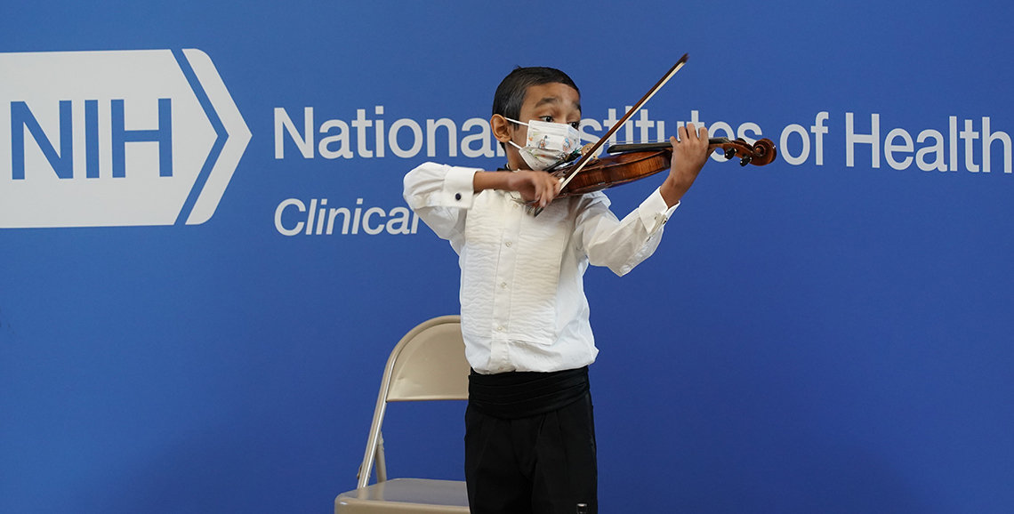 Caesar stands looking at his violin strings as he plays, in front of blue NIH banner in the atrium.