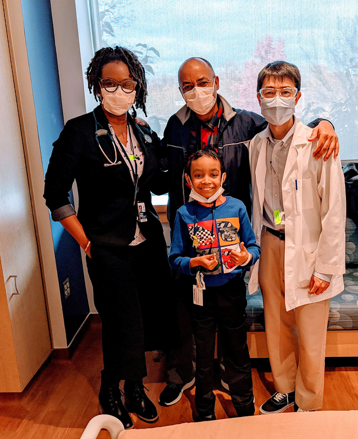 Caesar and his father pose with clinical staff--all masked, except Caesar whose mask is pulled down to reveal a wide smile. Many multi-colored leaves are seen through window behind them.