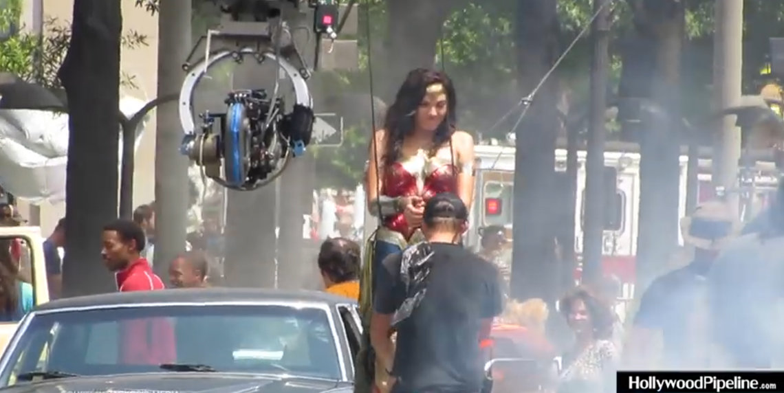 Flood, in a red jacket, is seen from side view, with movie star Gal Gadot, crew and camera boom and crane.