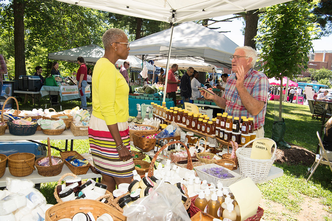Under tent, a black woman vendor stands behind tables of products and talks with white potential customer