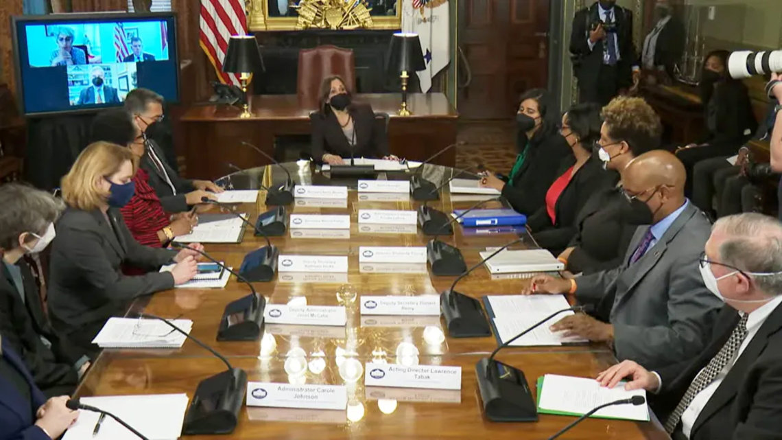 Men and women sitting around a long wooden table. All are masked, with long, white name cards in front of them. A video monitor with virtual attendees is displayed in the top left corner. 