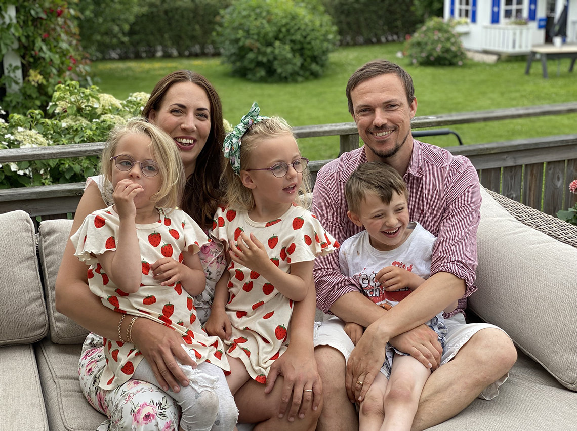 Isabella and Julia, in matching dresses with a strawberry pattern, and a laughing Hampus sit on their parents' laps on a couch in their backyard.