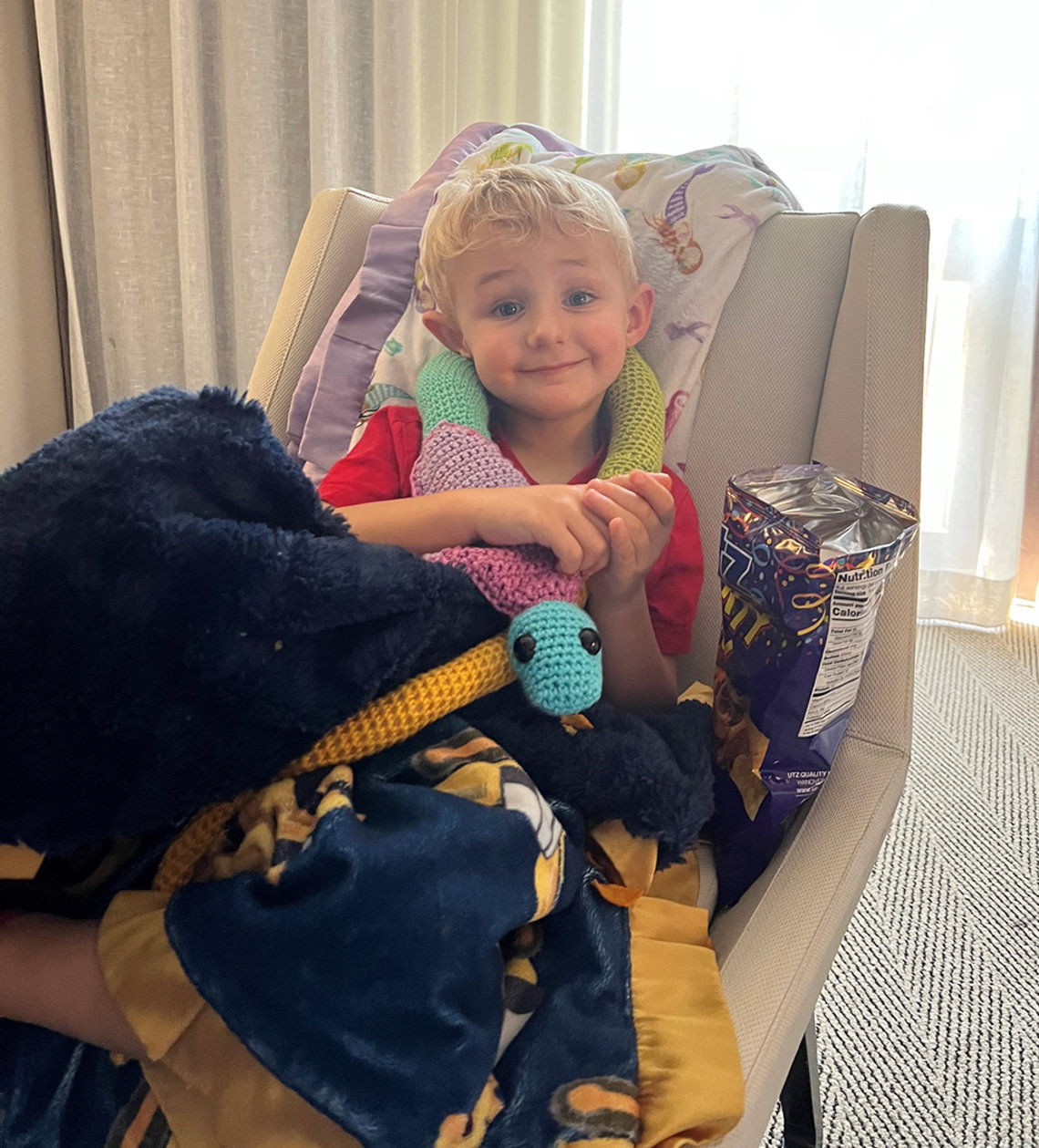 Grayson, age 3, sits in a chair with his blue and yellow snake plushie, his blanket and a bag of chips.