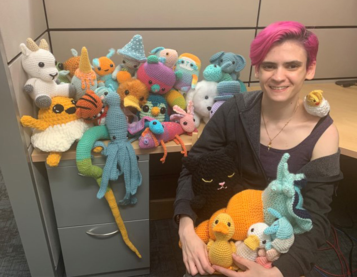 Dr. Head sits at a desk surrounded by 30 colorful plushies she crocheted.