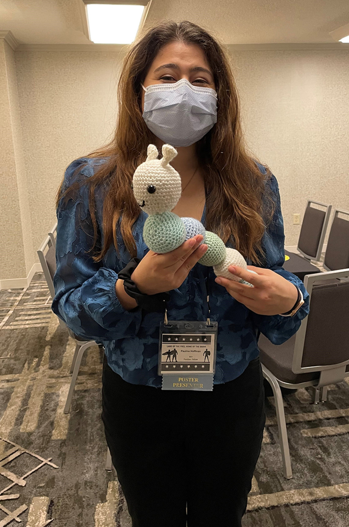 Hoffman, wearing mask, stands in conference room holding a blue, green and white caterpillar she crocheted.