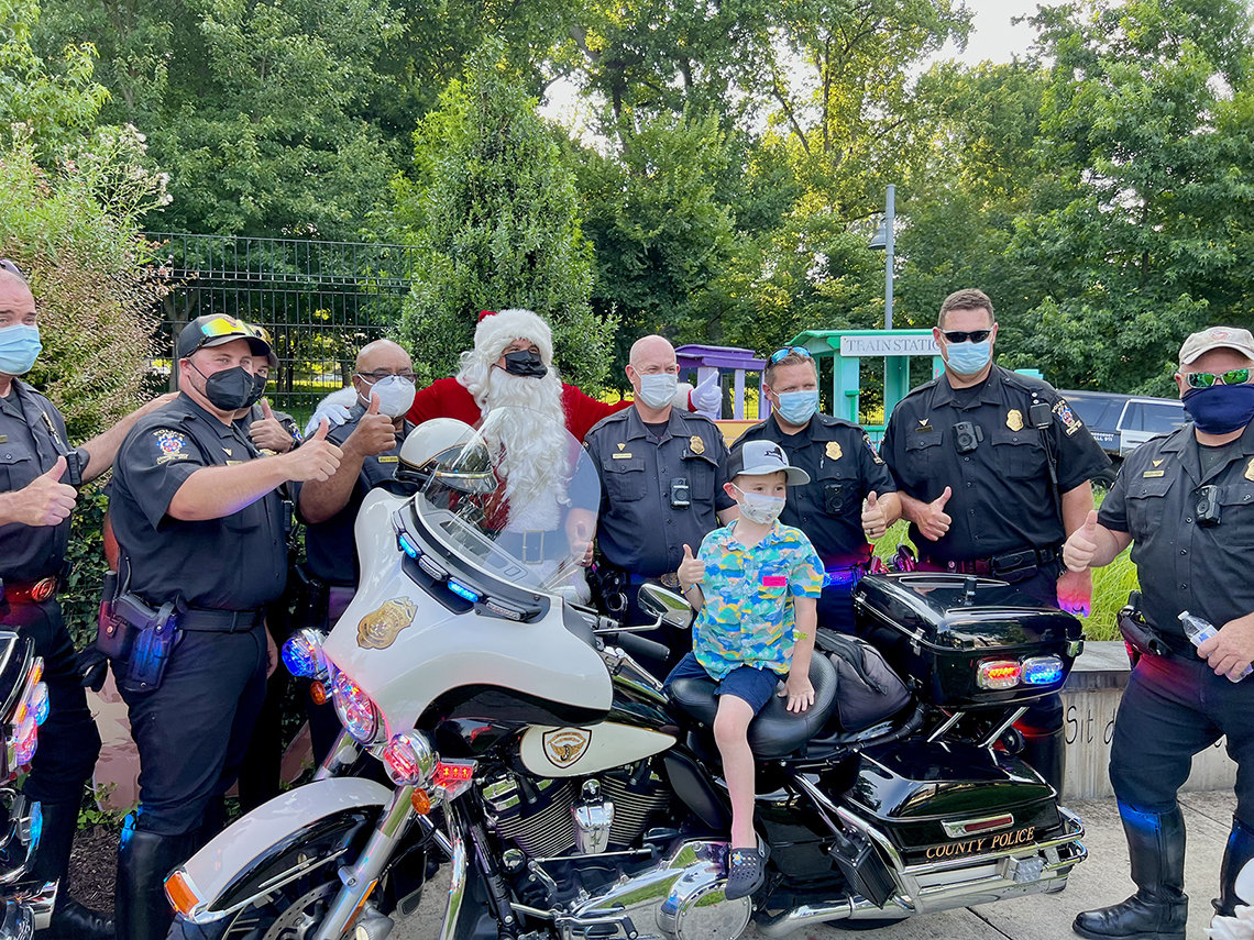 Child seated on motorcycle in front of uniformed police with Santa. All are masked and give the thumbs up sign.