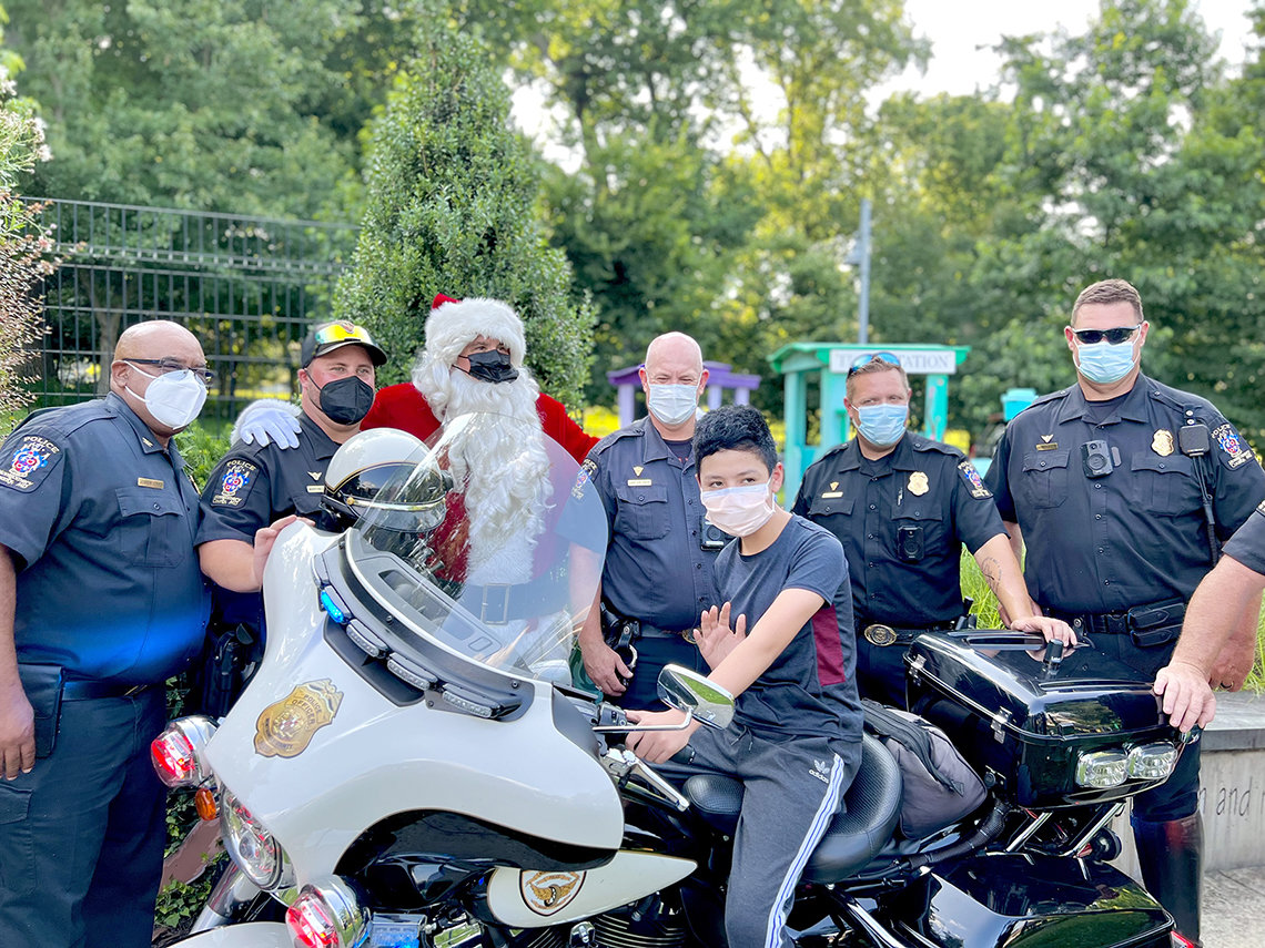 Child seated on motorcycle waves with uniformed police and Santa standing behind. All are masked.