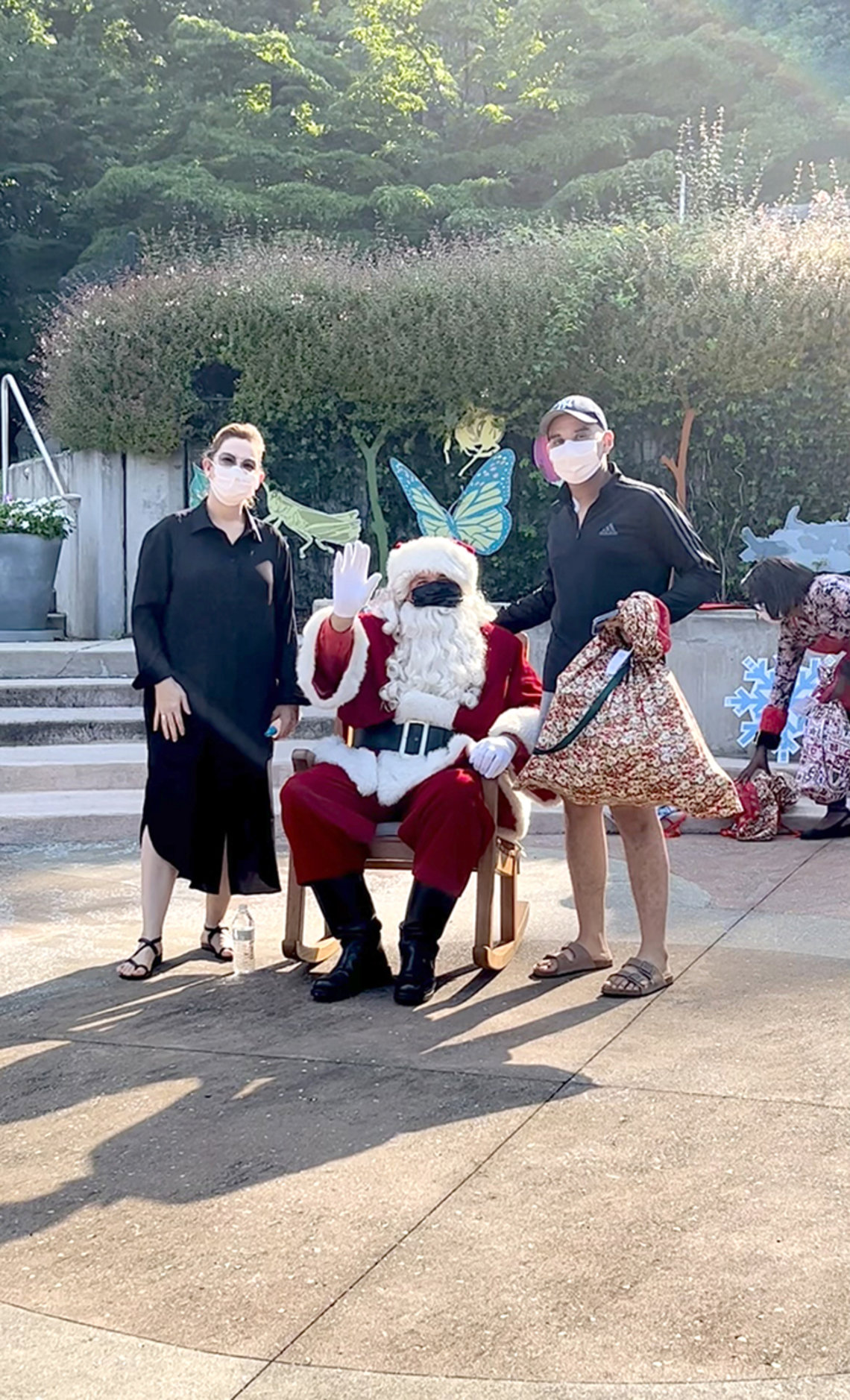 Masked Santa sits in a rocking chair with a masked woman and a masked man standing on either side