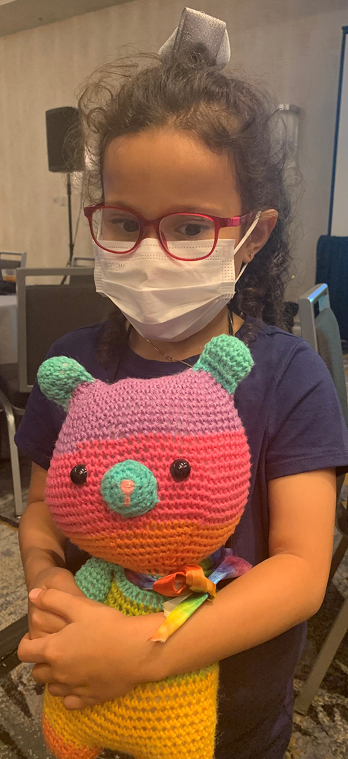 Catalina, in mask and red glasses, holds her colorful bear plushie.