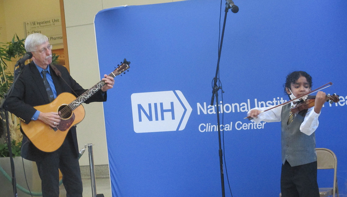 Collins plays guitar and Sant plays violin in front of blue NIH Clinical Center backdrop in the atrium.