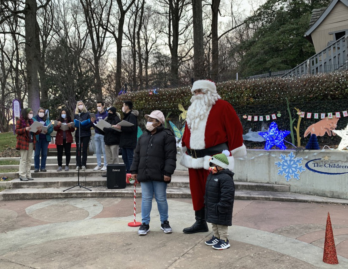 Santa stands with a young child and his mom, with 4 members of NIH's Nerds in Harmony singing into a microphone nearby.
