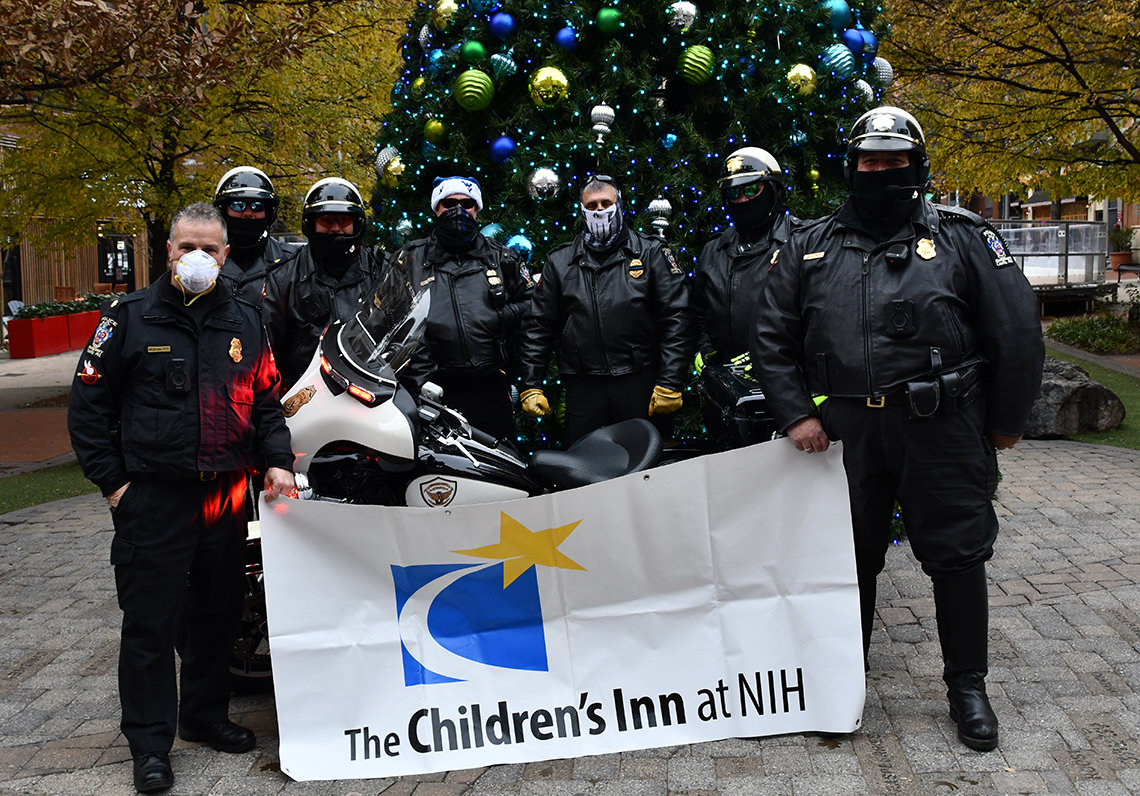 A group of officers holding a Children's Inn banner stand in front of the ice rink and giant Christmas tree in Rockville Town Square.