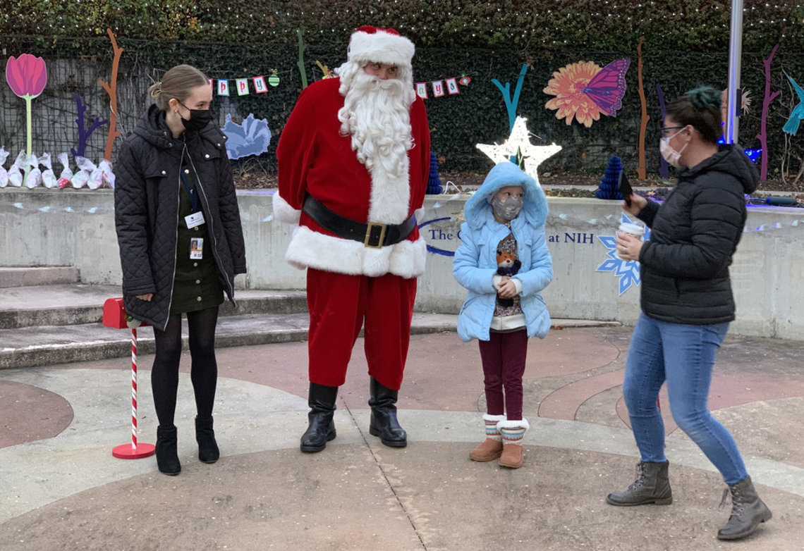 Santa stands next to a young girl and her mom and a rep from the Children's Inn, outside, with stars and flowers and other colorful decorations behind.