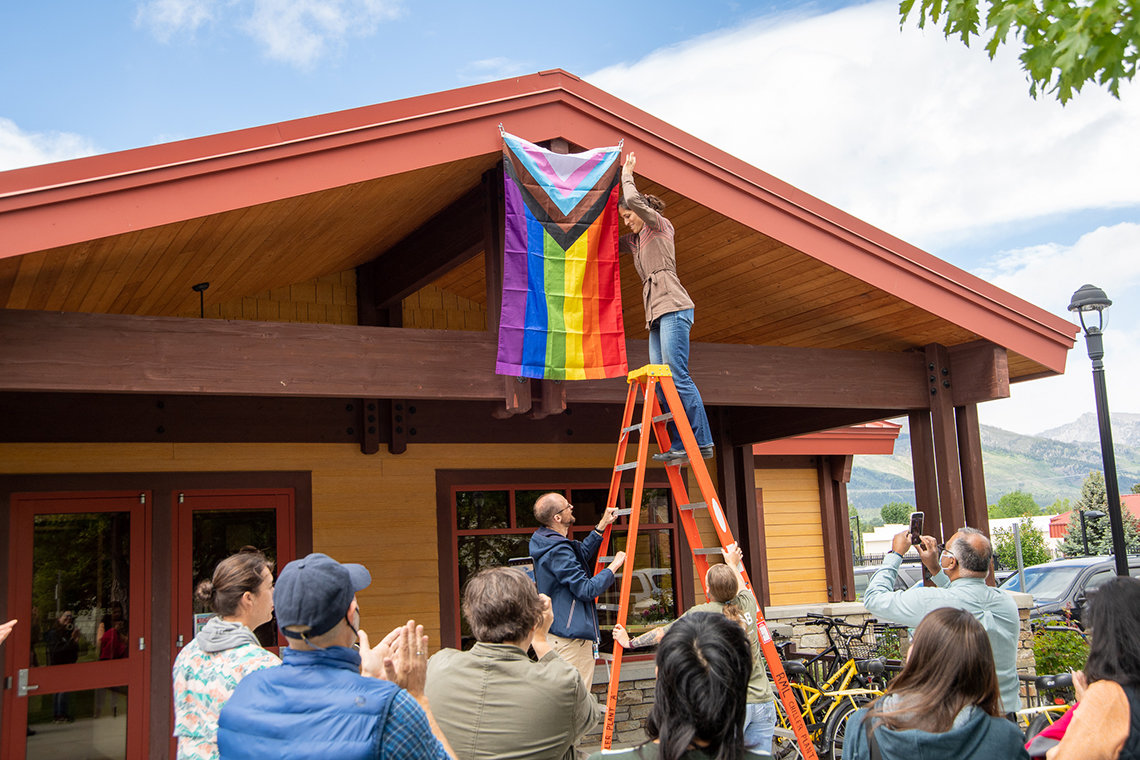 An employee stands on a ladder to hang the Progress Pride Flag on a building's awning while a group of employees look on