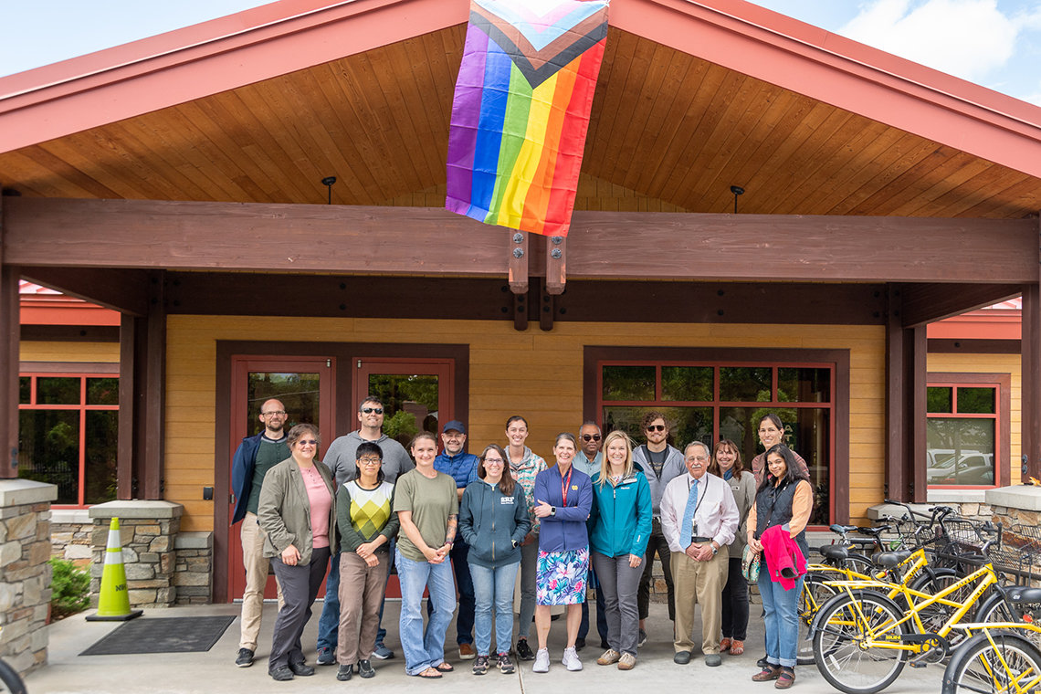 A group of RML employees stand in front of a building flying the Progress Pride Flag