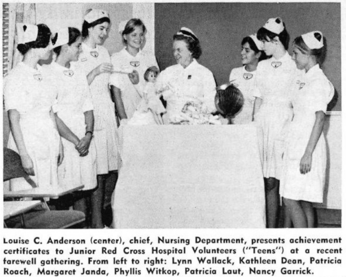 A group of young girls in retro hospital uniforms surround an older lady with a doll