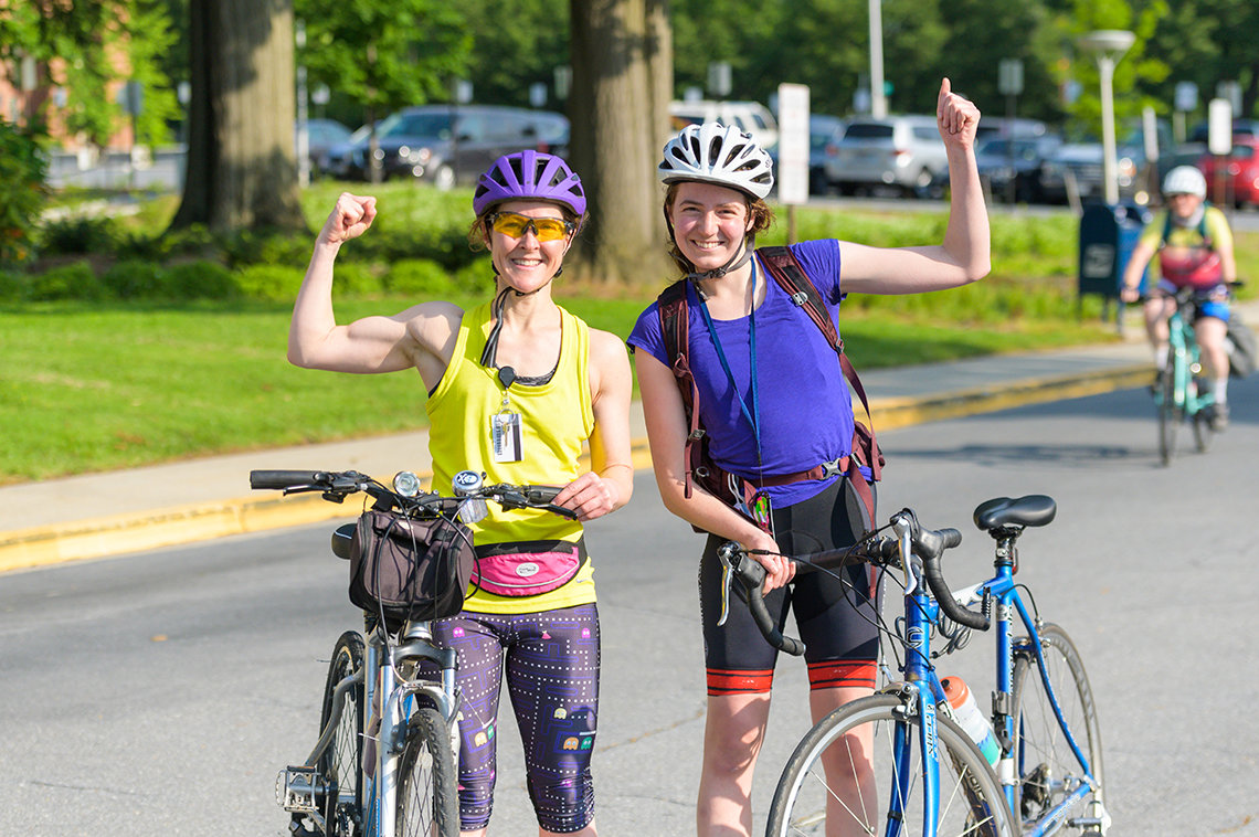 2 individuals in bike helmets hold bikes with one hand while flexing muscles and giving thumbs up with the other hand