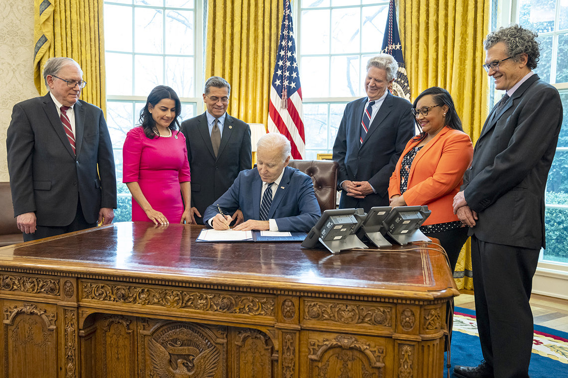 President Biden seated at desk with several people looking over his shoulder