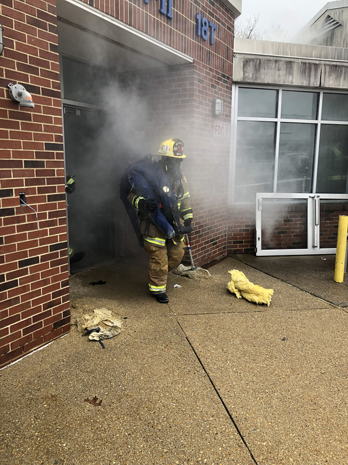 Surrounded by smoke, firefighter in full gear and helmet exits building with dummy on shoulder