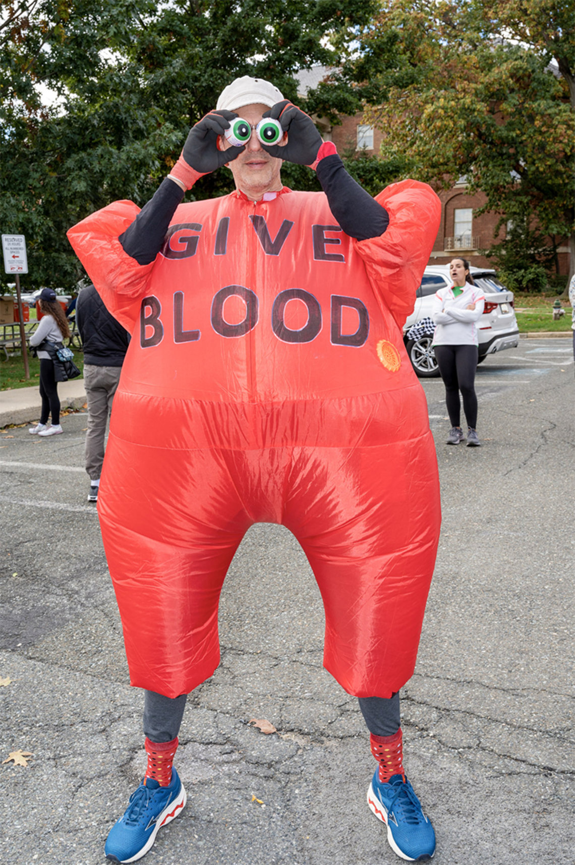 Hayes in red, inflatable costume that reads "Give Blood," holds 2 plastic eyeballs over his eyes outside, in front of Bldg. 1.