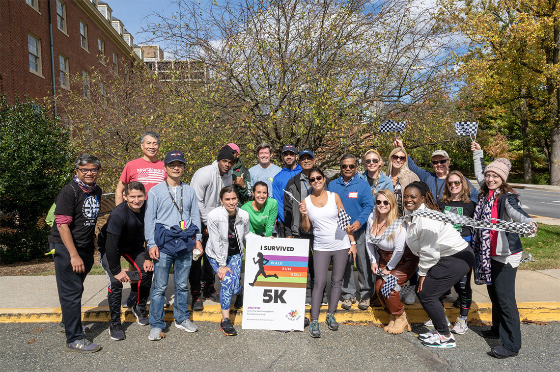 A group of smiling people gather around sign that reads "I survived Walk/Run/Roll 5K outside on the NIH campus.