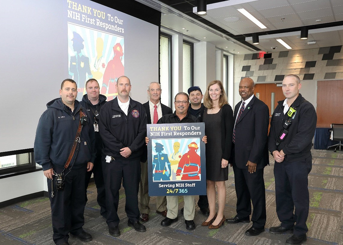 Large group of people stand behind a poster with a graphic representing a firefighter, EMT, and police