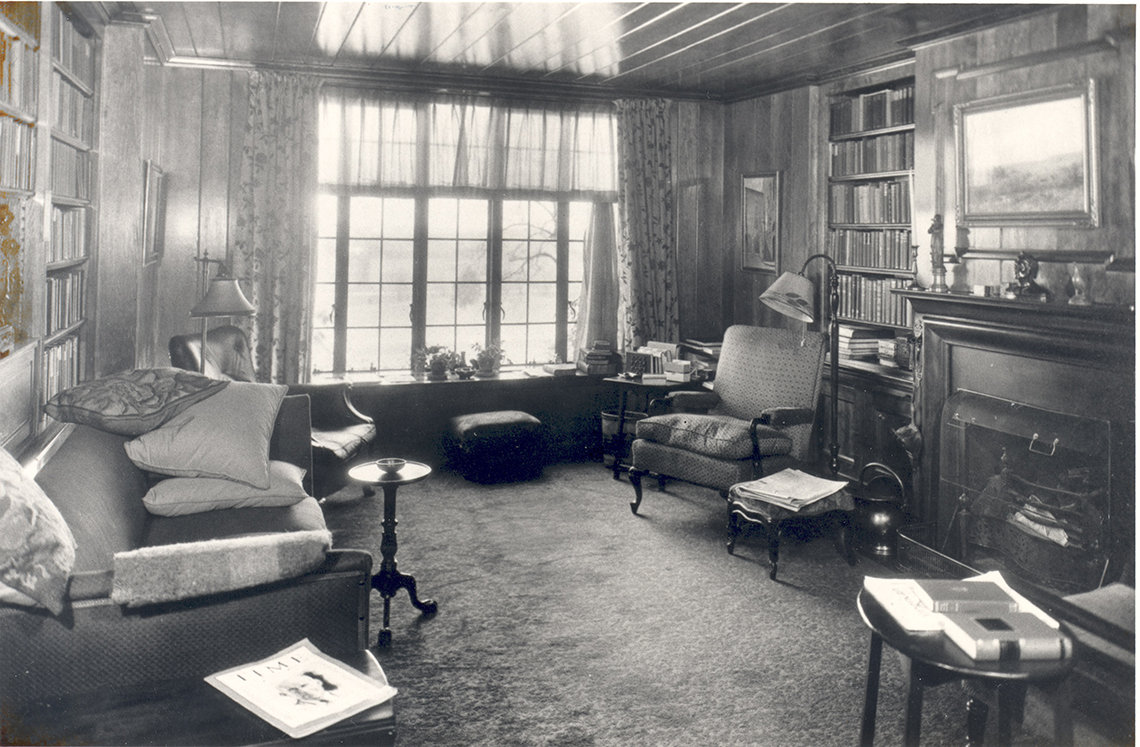 A black and white photo of the library when the Wilson's lived there in the 1930's.The wood paneled room has a fireplace and several pieces of furniture, including chairs, side tables and a couch. 