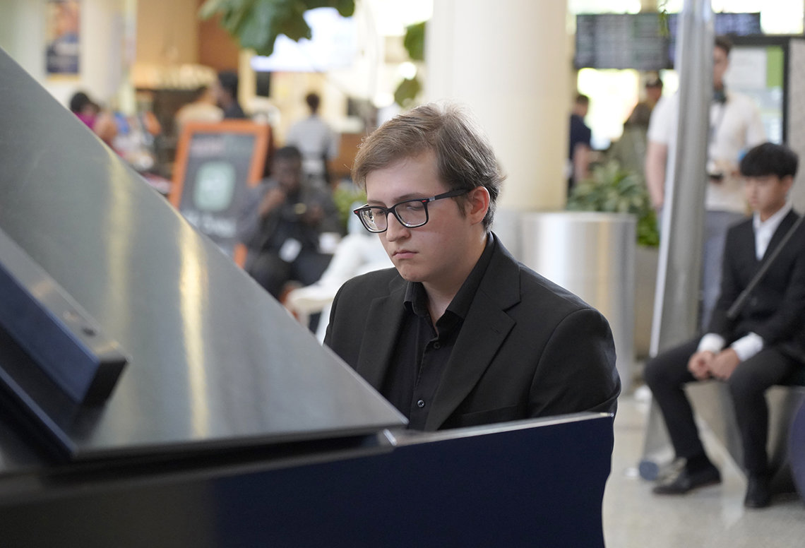 A young man in a black suit sits at the piano in the CC atrium.