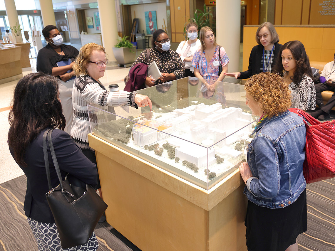 Group of adults gather around an architectural model of the hospital, as a woman points to features of the structure