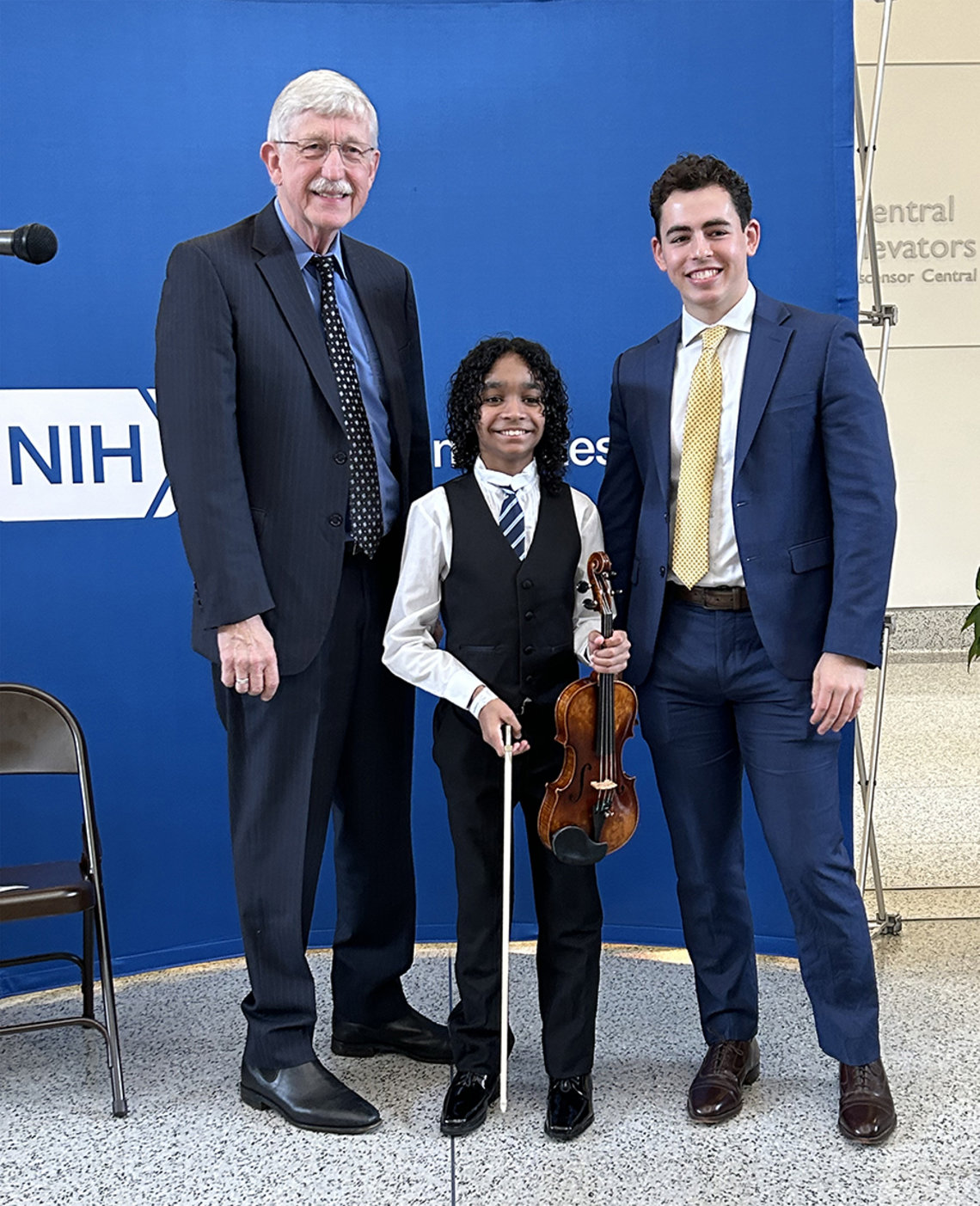 Collins, Caesar (holding his violin) and Masi stand together for a photo in front of a blue backdrop