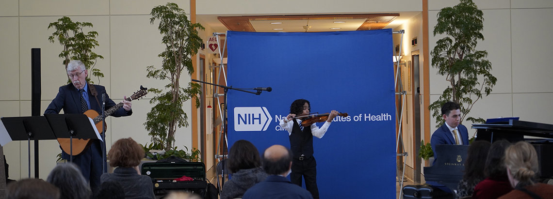 Wide shot shows Sant on violin, Collins on guitar and Masi on piano in the Clinical Center atrium.