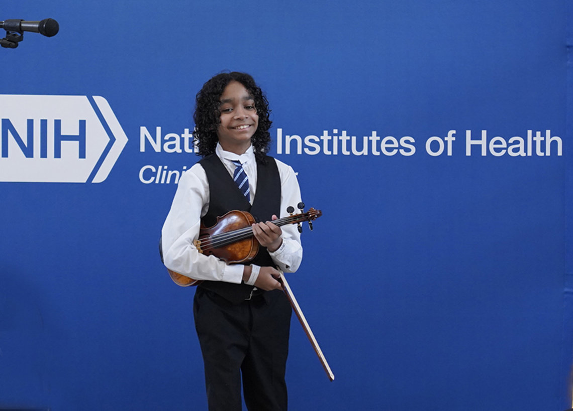 Smiling, Sant holds his violin and bow standing in front of blue NIH banner.