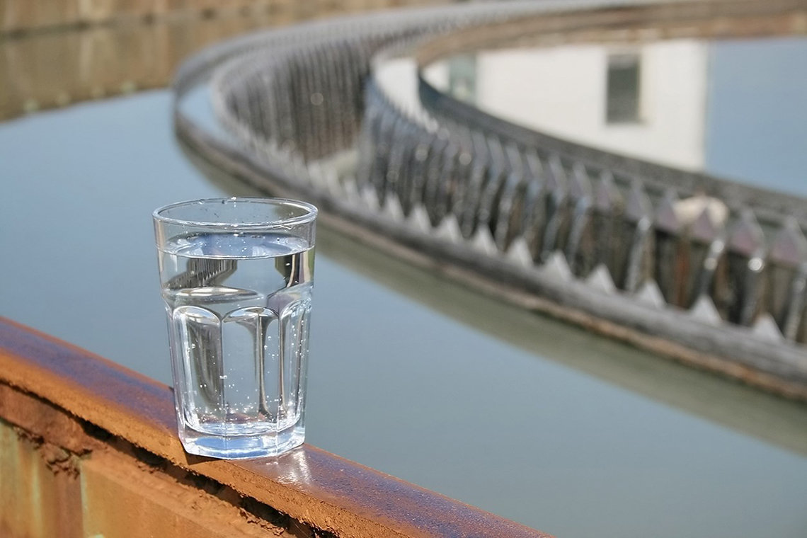 A clear glass of water balances on a rusty rail near a body of water