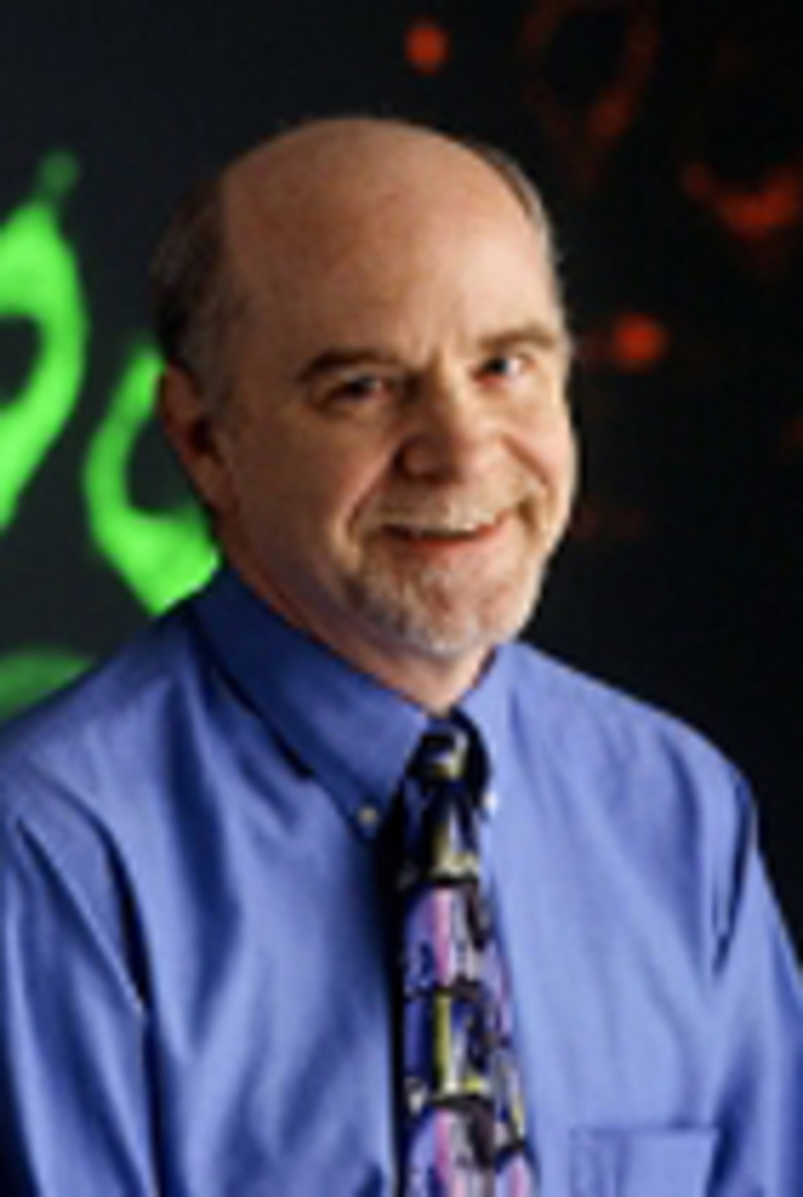 Dr. O'Shea, in a blue shirt, poses against a black and green background