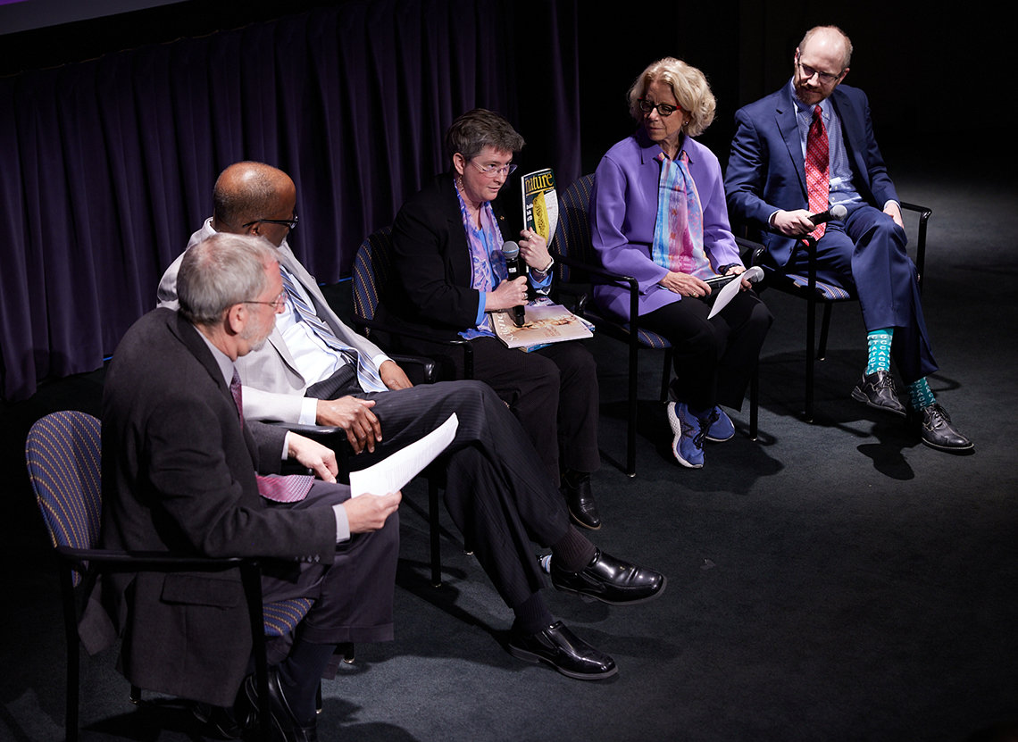 A panel sits on stage, with Dr. Rutter in the middle holding up a copy of Science magazine.