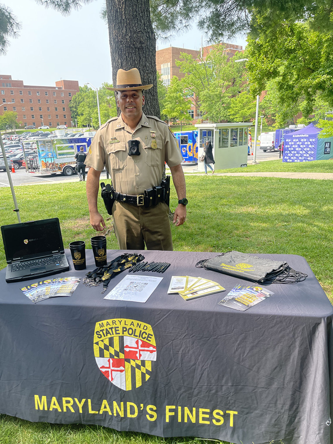 A smiling state trooper stands by info table, with a table covering that reads: Maryland's Finest.