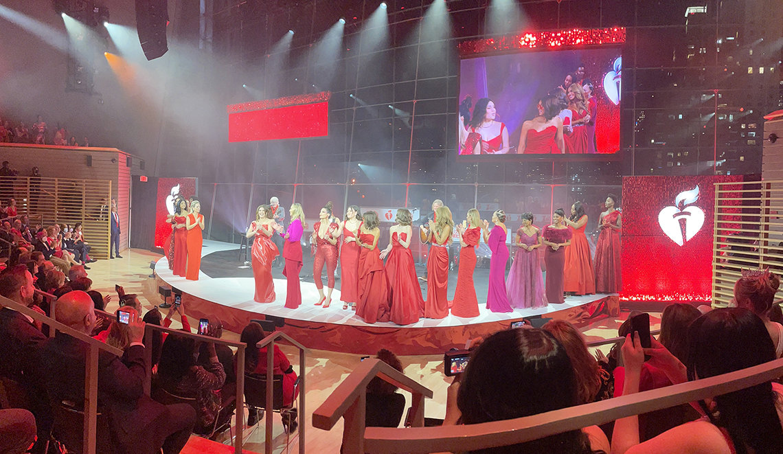 Women in red dresses stand on circular stage beneath lights as audience watches.