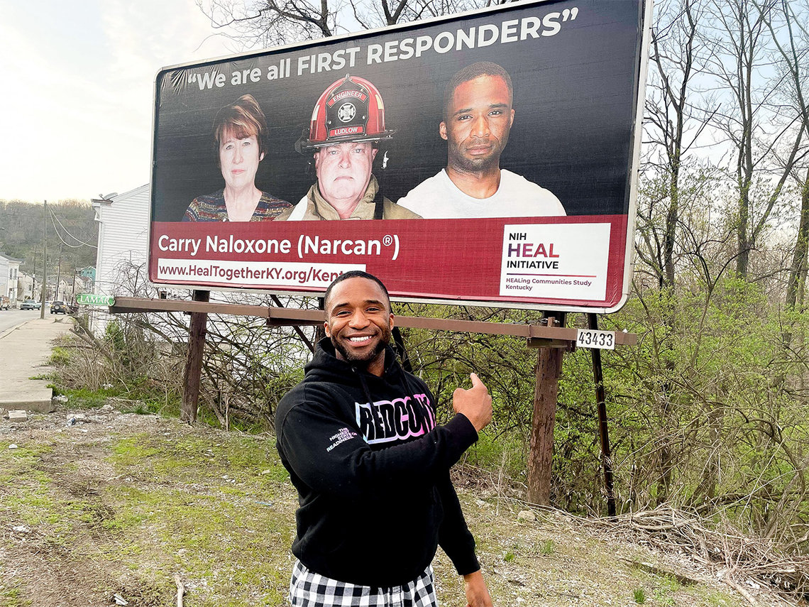 McCarty stands pointing to himself on billboard behind him, that reads NIH HEAL initiative