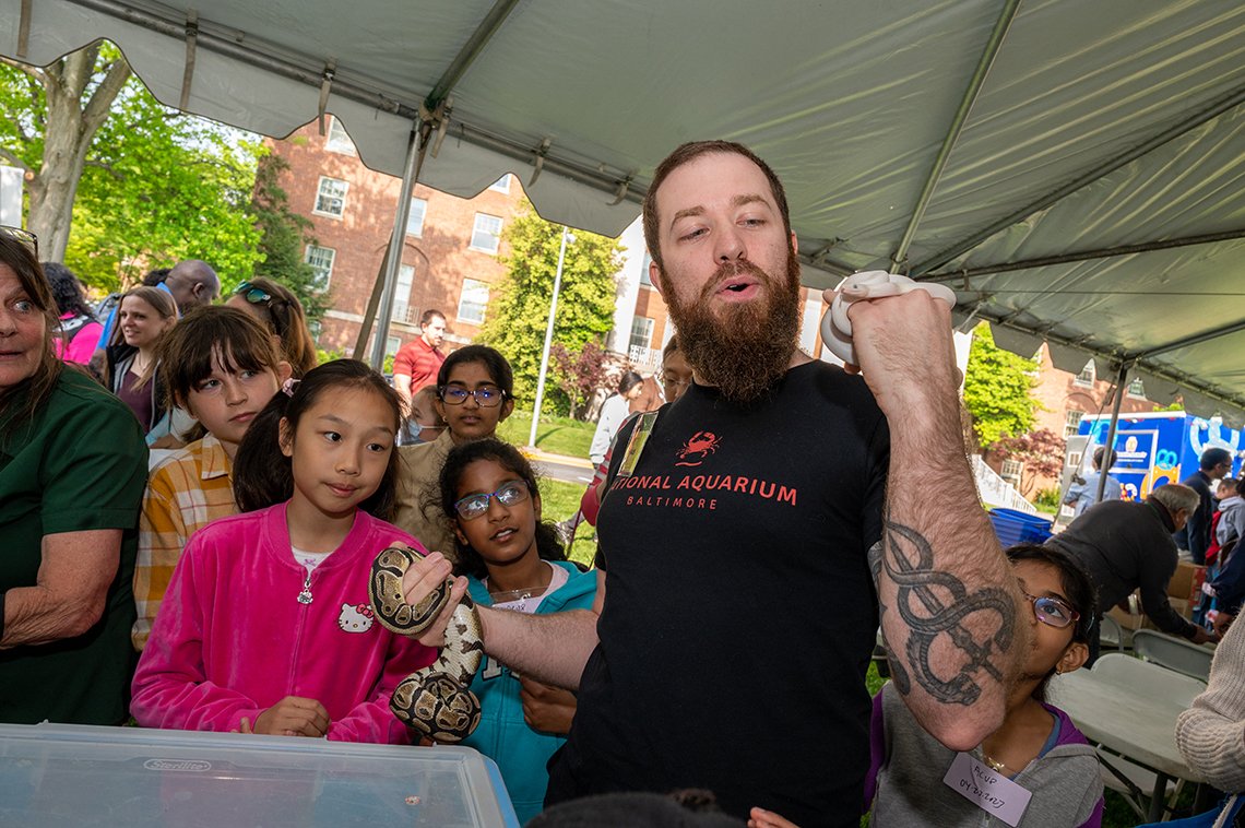 Man with snake tattoo holds up two live snakes for crowd of children.
