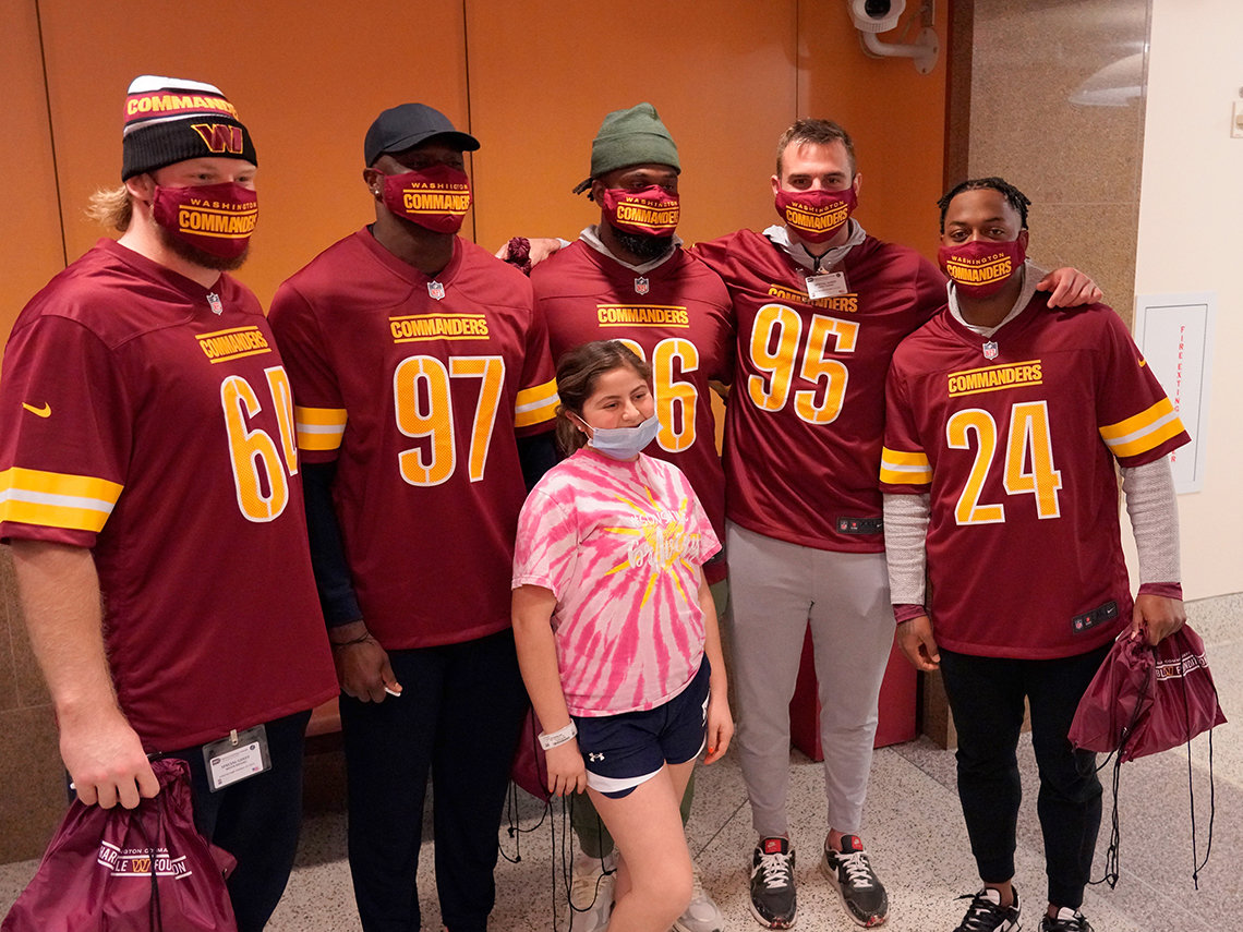 Female youngster in tie-dyed pink T-shirt smiles behind face mask with five men in face masks and football jerseys behind her.