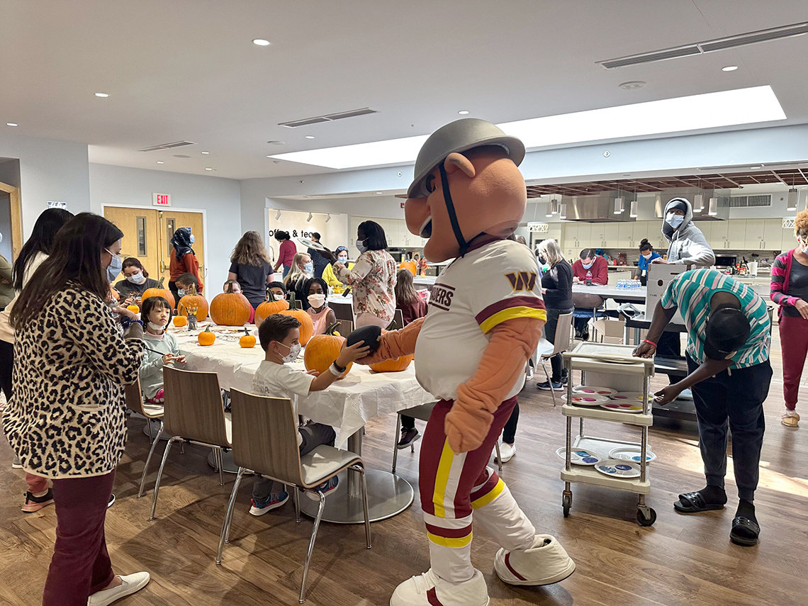 Person in oversized pig costume with combat helmet and Commanders jersey reaches toward youngster seated at craft table.