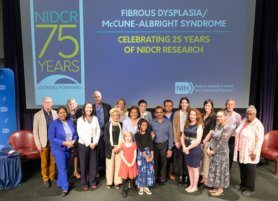 A large group of staff and patients stand on stage in front of slide: NIDCR 75 years - Fibrous Dysplasia-McCune-Albright Syndrome
