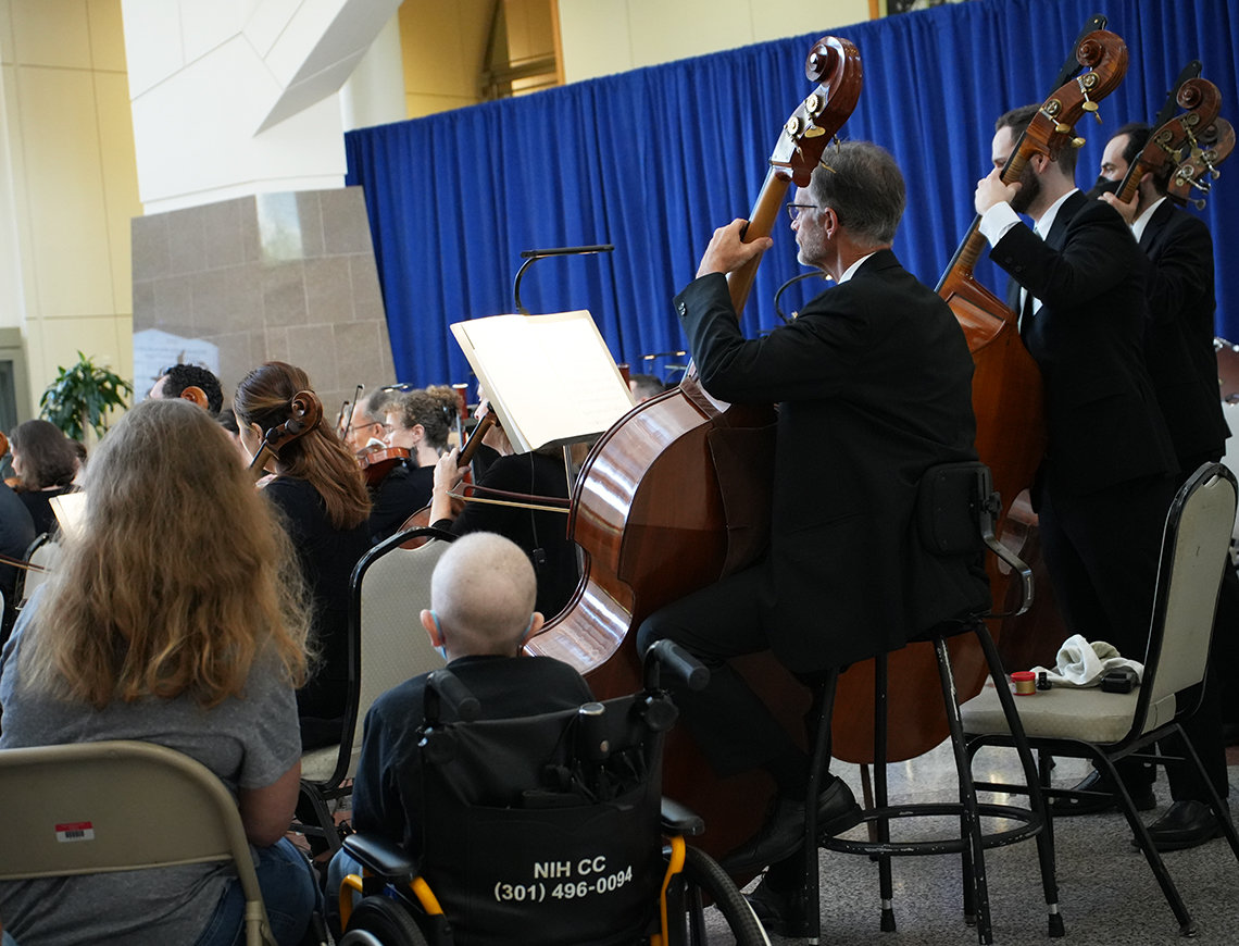 Viewed from behind, a child sits in a wheelchair next to 3 cellists