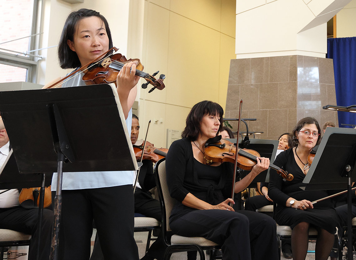 Qiao holds bow to violin, with string section behind her in the atrium