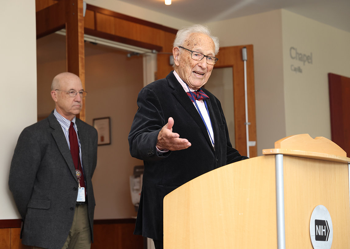 Arias in suit and bowtie gestures at the podium as Gilman watches from behind