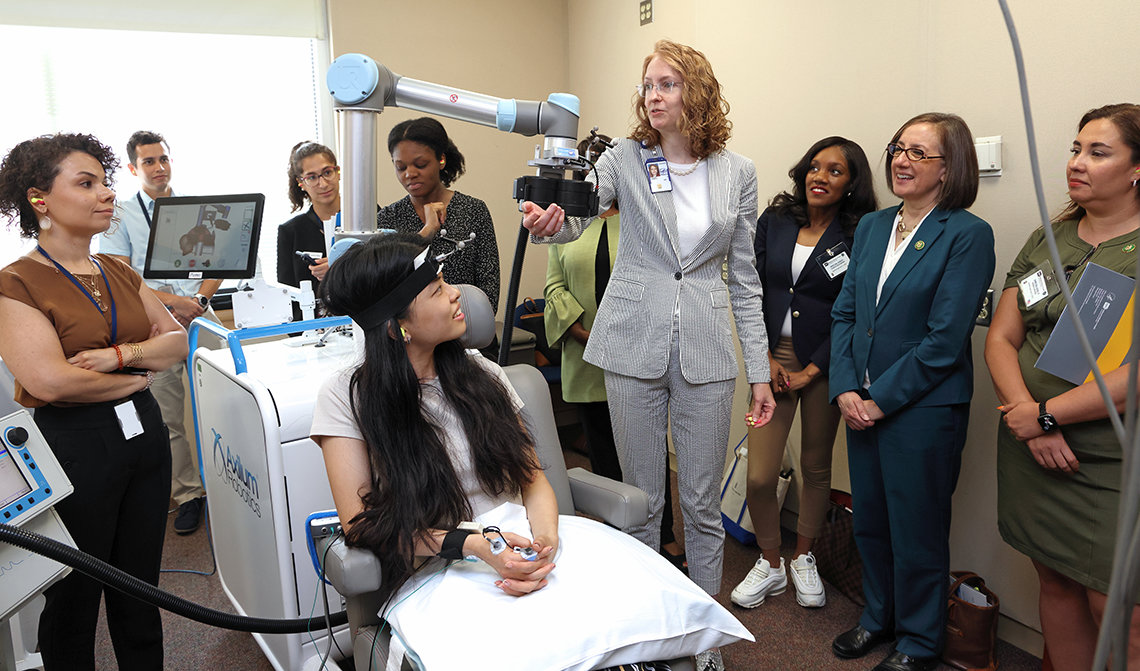 A volunteer in chair with headband and wires, hooked up to noninvasive magnetic stimulation machine, guided by Lisanby, as congresswomen and staffers watch demonstration