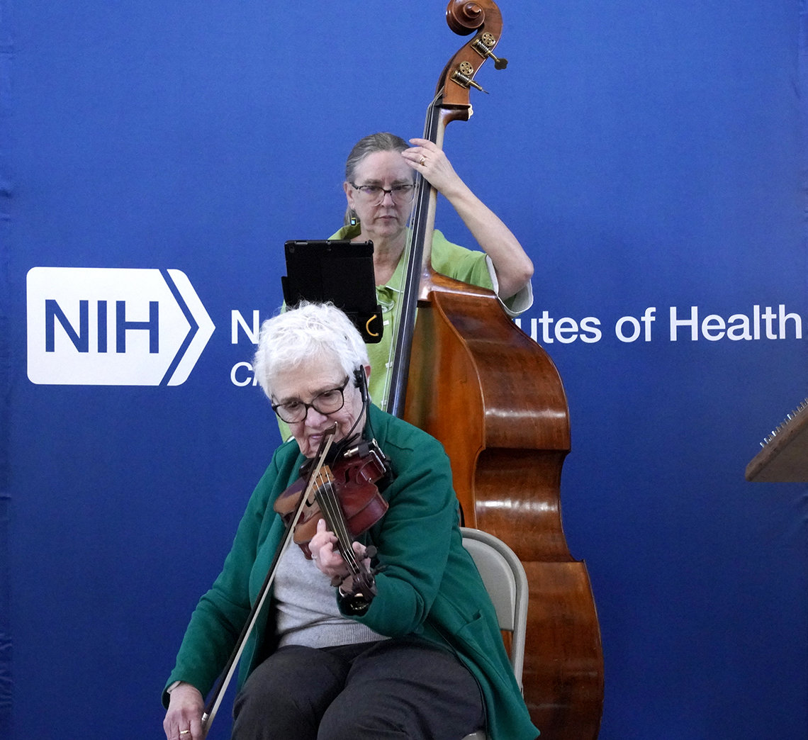 A seated woman plays the violin. Behind her, a standing woman plays the bass.