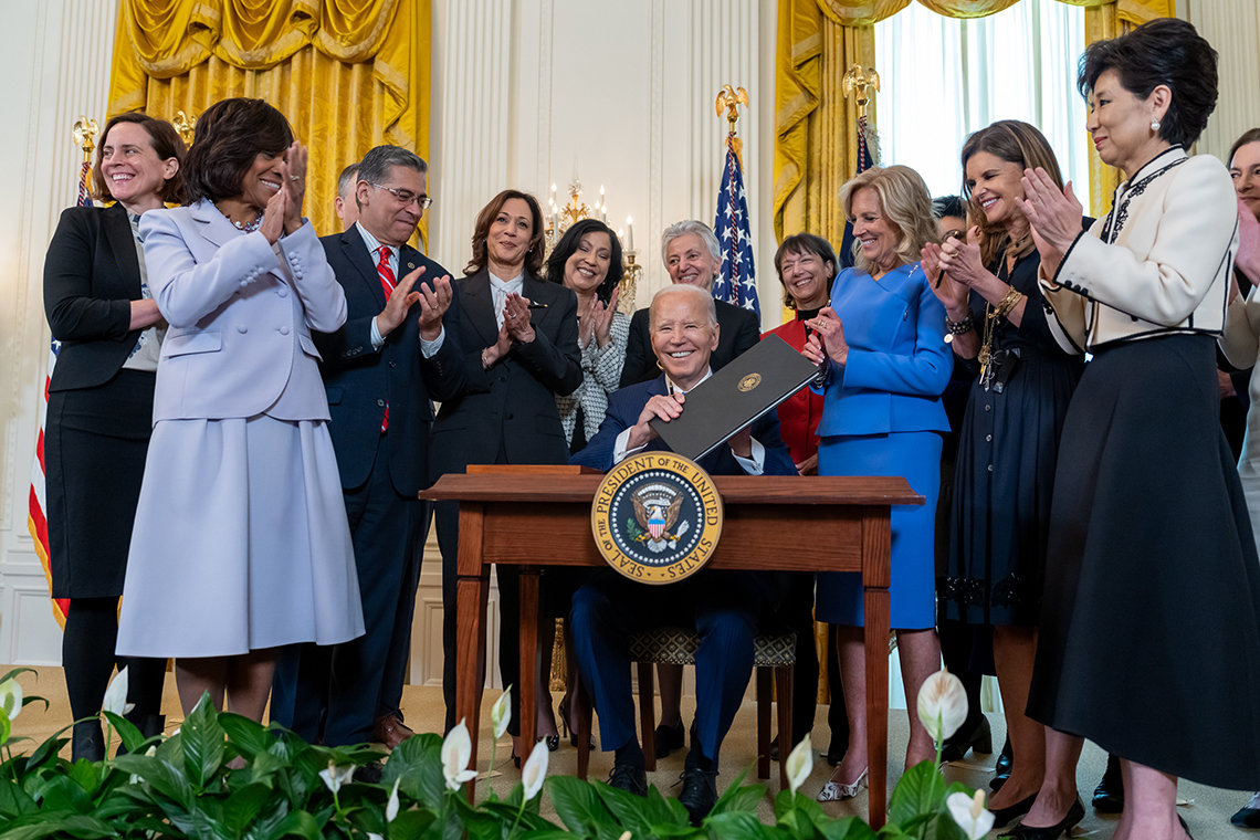The HHS secretary and NIH leaders gather around a smiling President Biden, seated at Oval Office desk, as he holds binder with signed executive order.