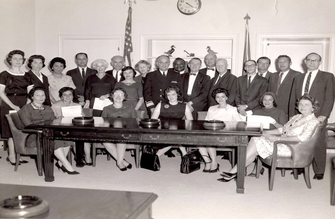 24 people, some seated at a conference table, gather for a group picture